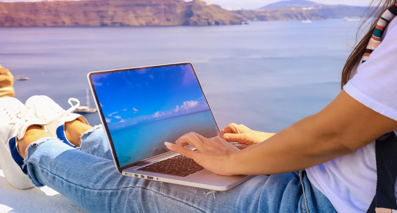 Person wearing jeans working on a laptop with ocean and cliffs in the background on a sunny day.