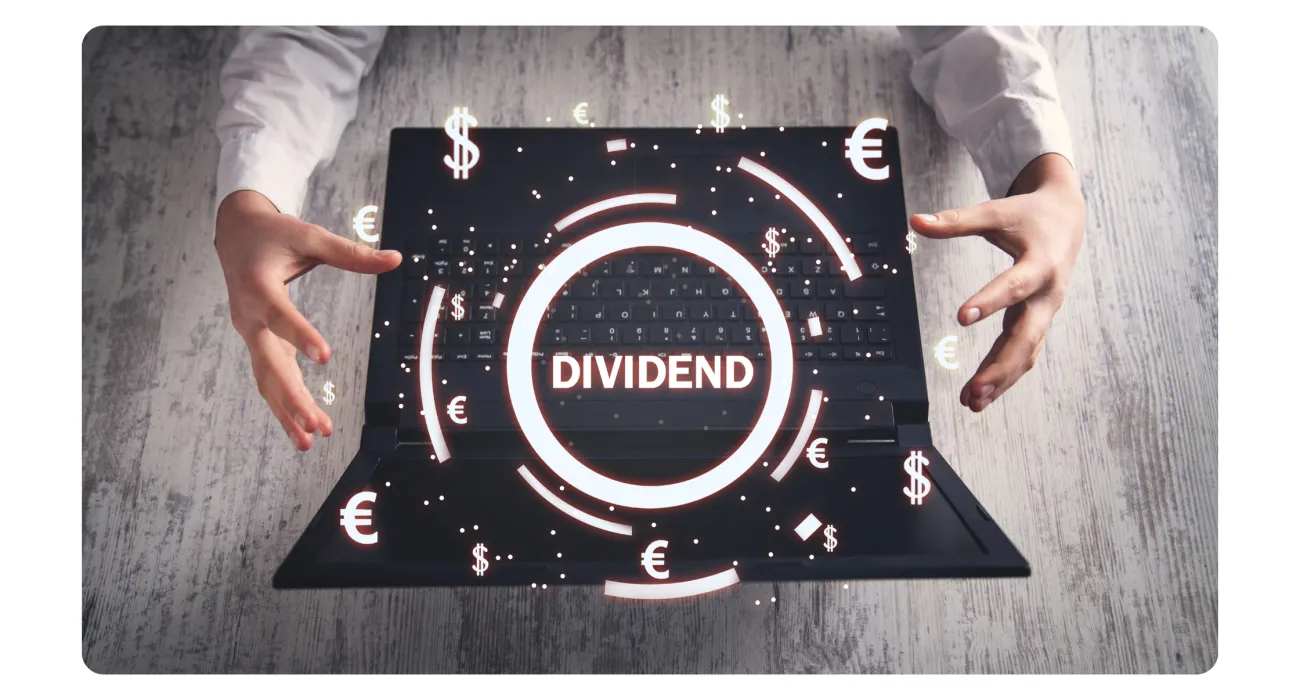 Hands holding a laptop with glowing dividend text and floating dollar and euro currency symbols over keyboard.