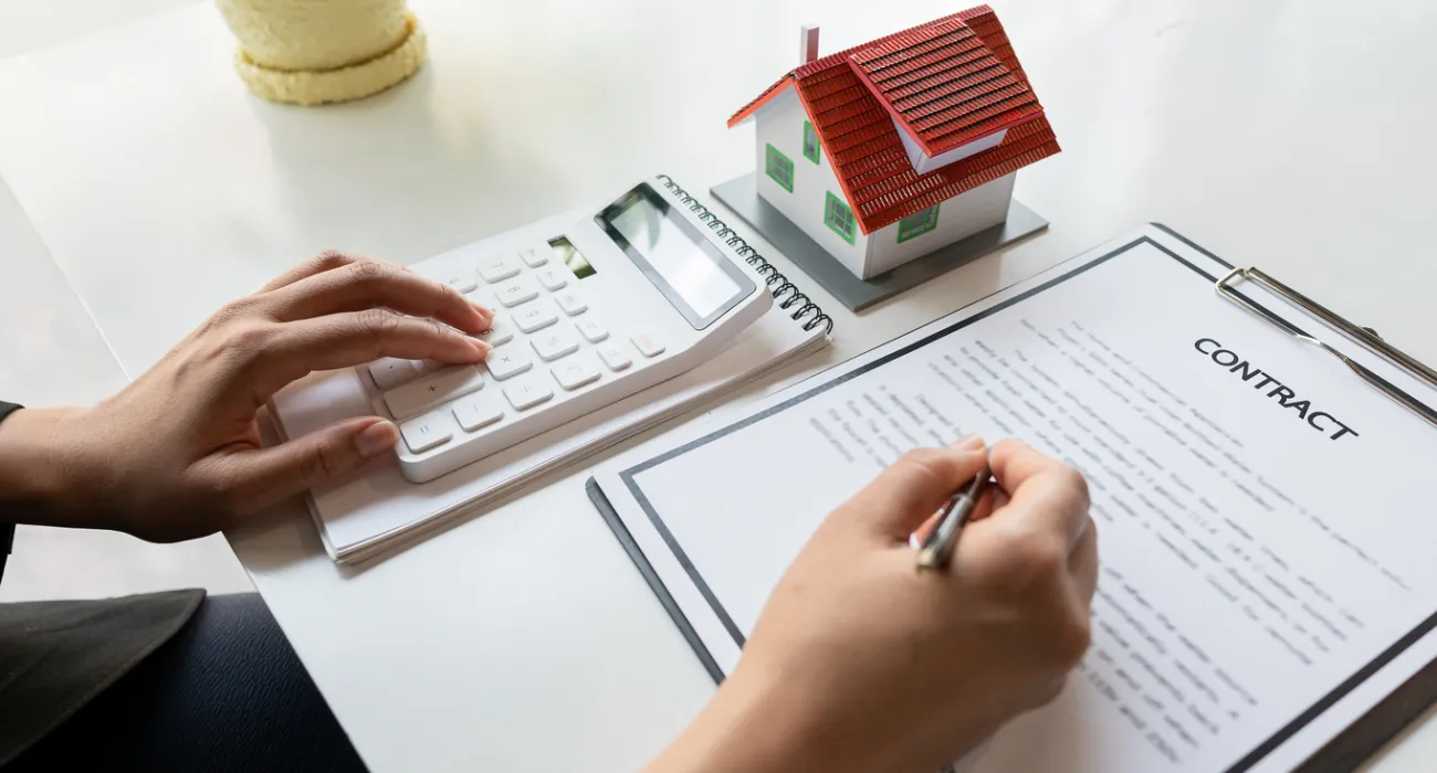 Person calculating costs for a property contract with a miniature house and document on the table.