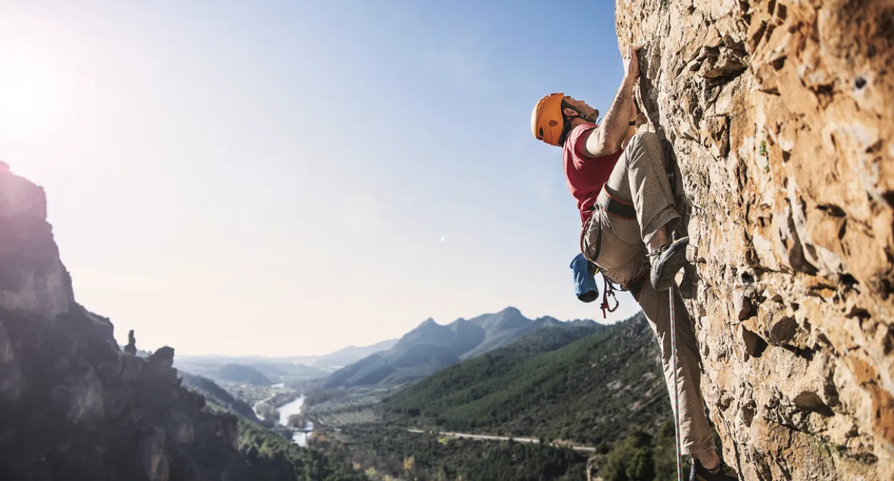 Man climbing a steep rocky cliff with safety gear against a mountain valley backdrop in bright daylight