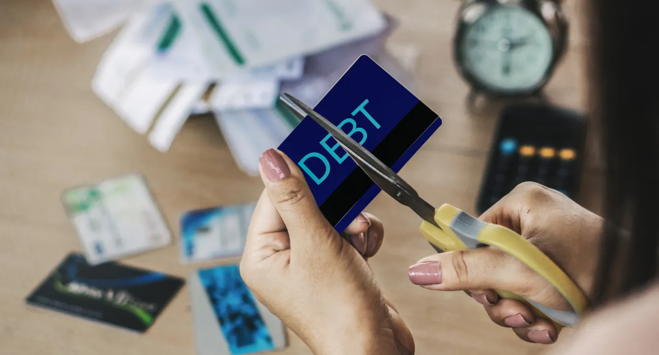Person cutting a credit card labeled debt with scissors, symbolizing debt reduction or financial freedom.