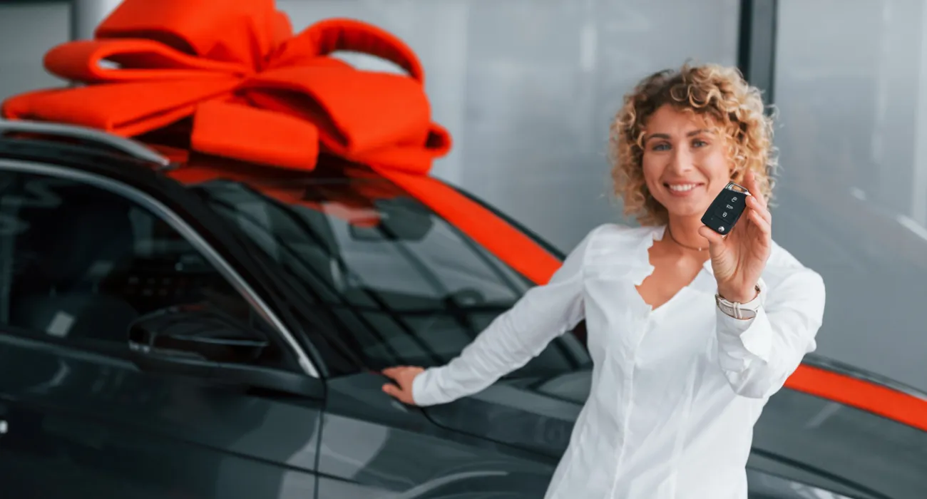 Smiling woman in white shirt holding car key next to a black car with a large red bow on the roof.