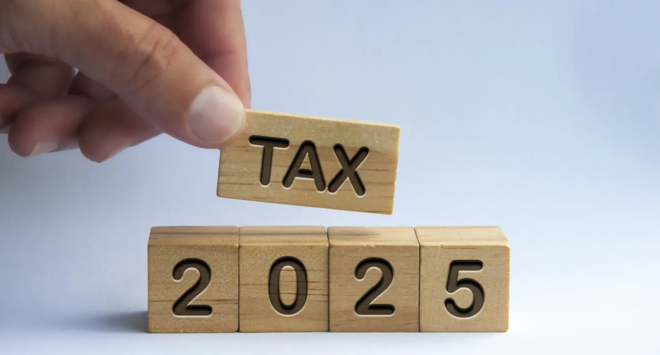 Hand holding a wooden block with 'TAX' above wooden blocks spelling '2025' on a white background.
