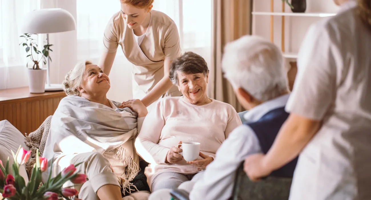 Senior adults enjoying tea and friendly care in a bright living room setting with attentive caregivers