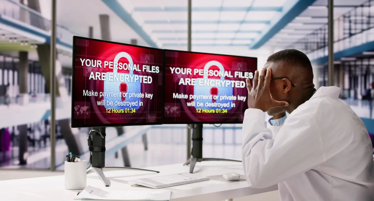 Man in white shirt shocked at dual monitor ransomware message demanding payment to decrypt files with countdown timer.
