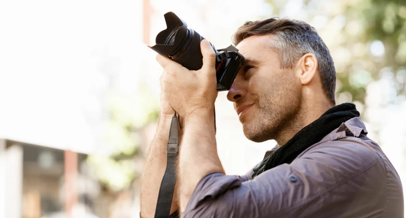 Man with grey hair focusing while taking a photo with a professional camera outdoors on a sunny day.
