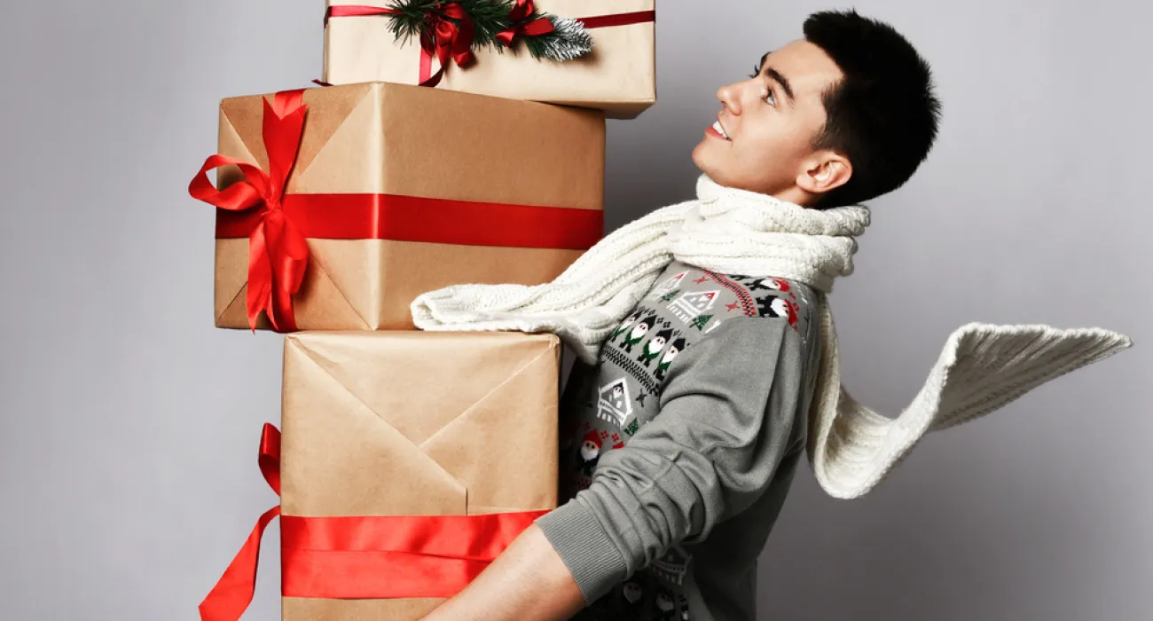 Young man in holiday sweater and scarf balancing multiple wrapped Christmas gifts with red ribbons.