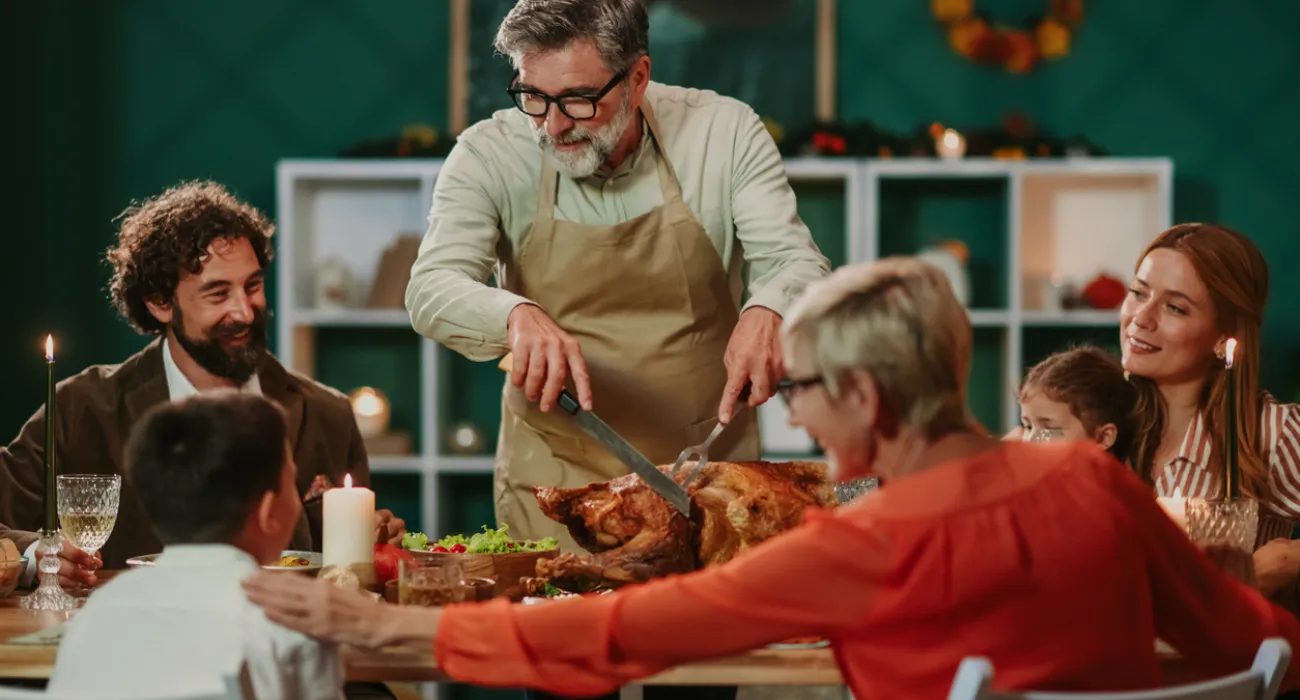Family gathered around dining table as man carves roasted turkey during festive meal together.