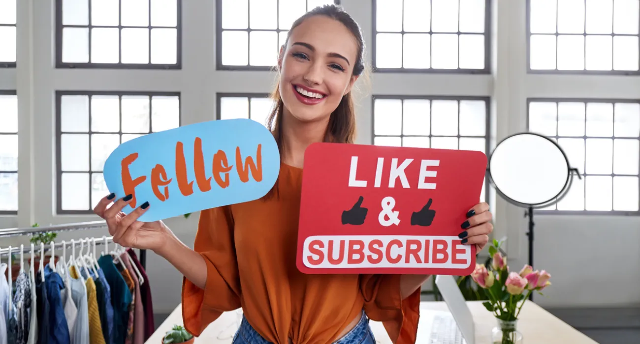Smiling woman holds follow and like & subscribe signs in a bright modern studio with clothing rack and flowers.