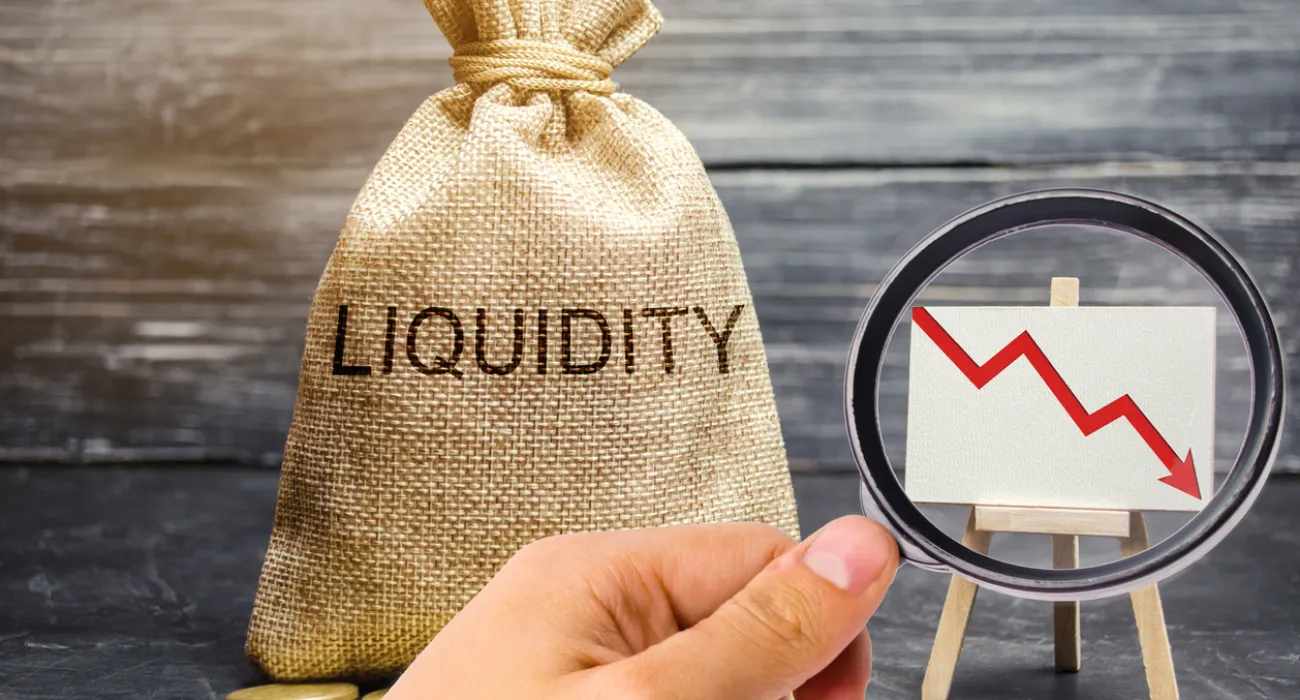 Hand holding magnifying glass focused on downward red graph beside money bag labeled liquidity and coins