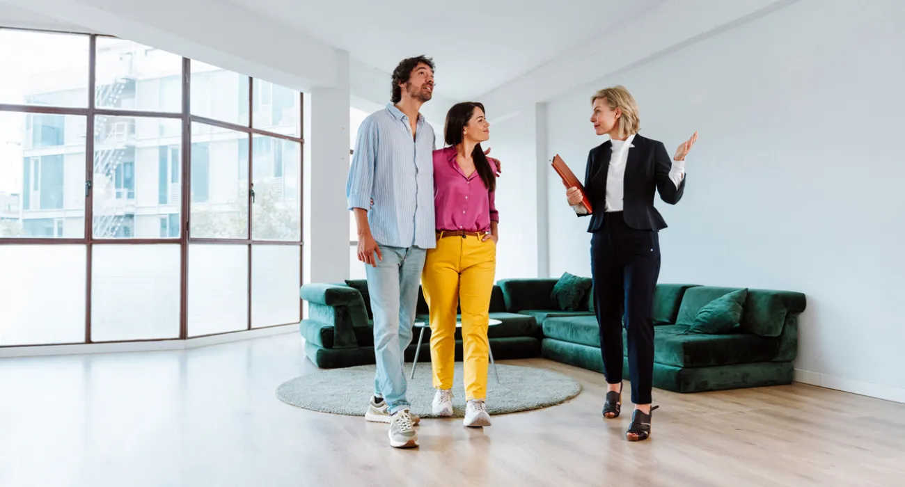 Real estate agent showing a couple a bright, modern living room with large windows and green sofa.