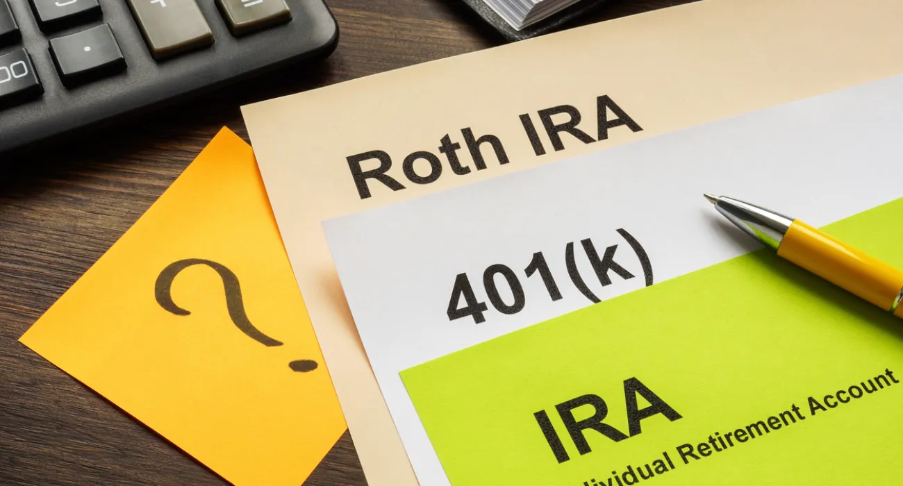 Documents labeled Roth IRA, 401(k), IRA with pen and calculator on wooden desk indicating retirement planning.