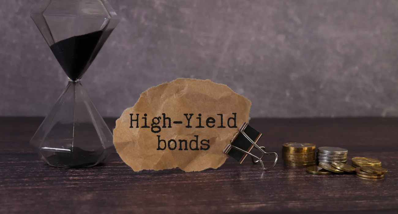 Hourglass, torn paper saying High-Yield bonds, and stacks of coins on wooden surface representing investment concept