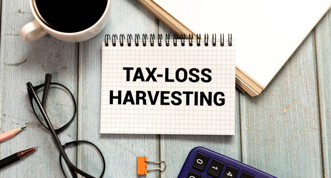 Notebook with Tax-Loss Harvesting text surrounded by coffee cup, glasses, calculator, pencils, and paper on wood table.