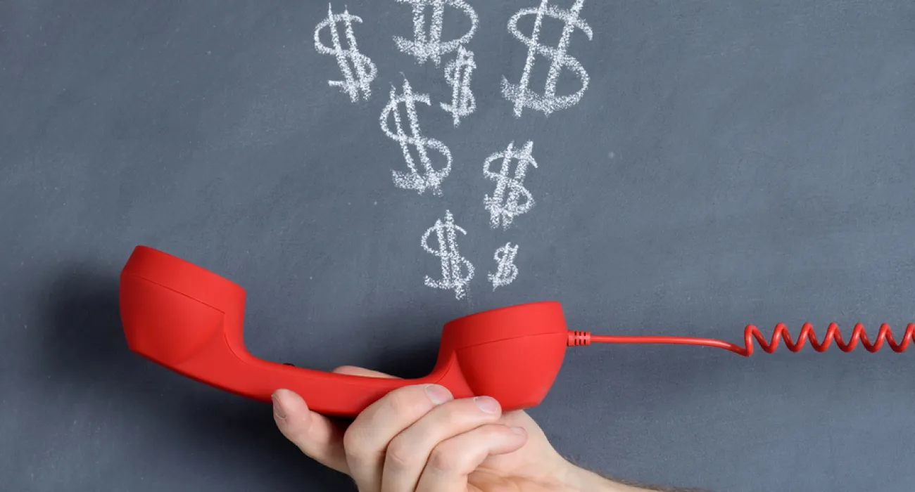 Hand holding red phone receiver with dollar signs drawn above it on chalkboard background.