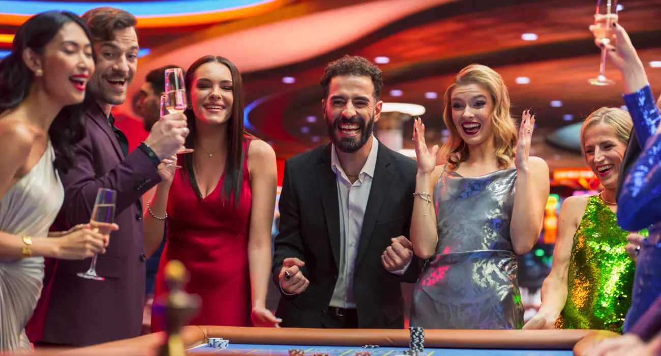 Group of stylish people celebrating and cheering around a casino gambling table with drinks in hand