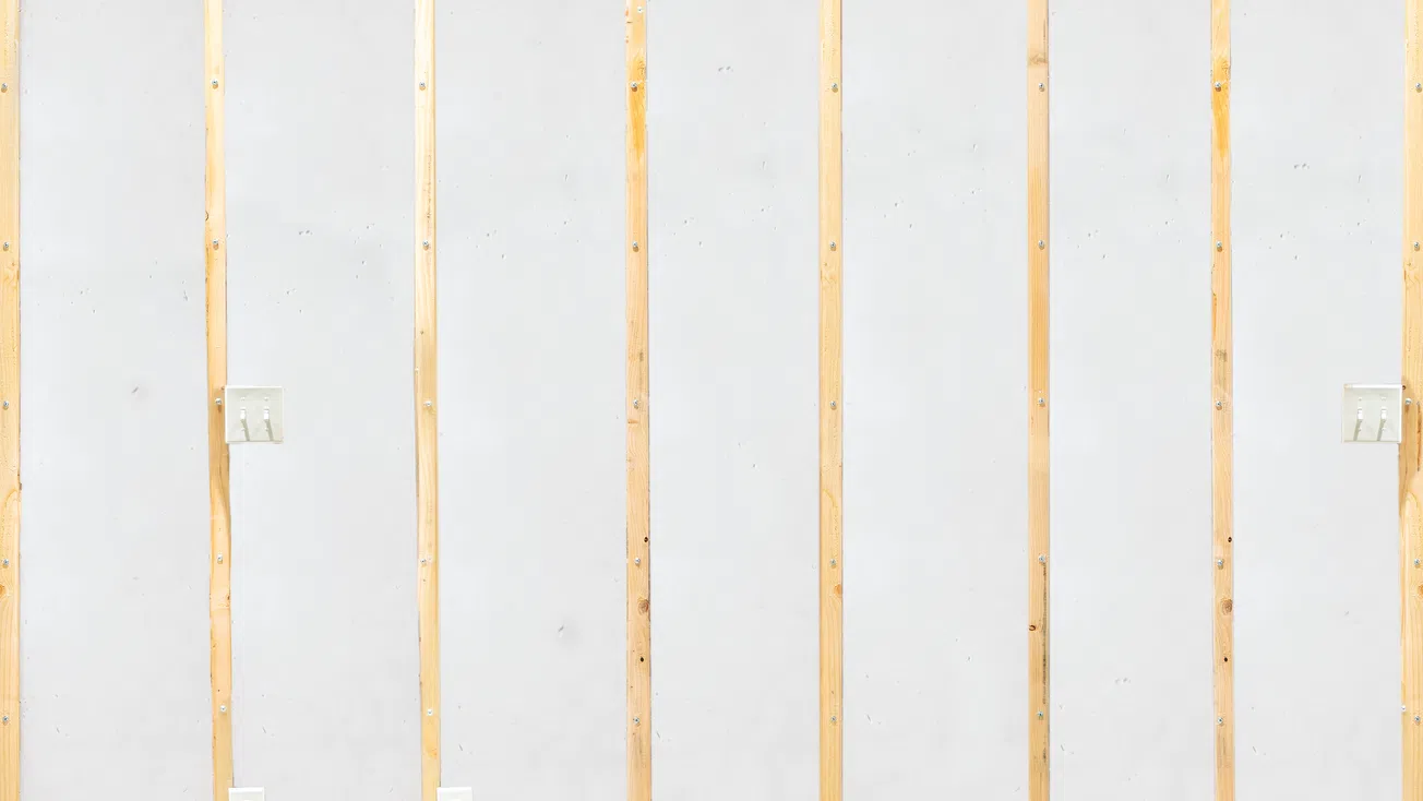 Unfinished wall with exposed wooden studs, drywall panels, electrical outlets, and light switches in a construction setting.