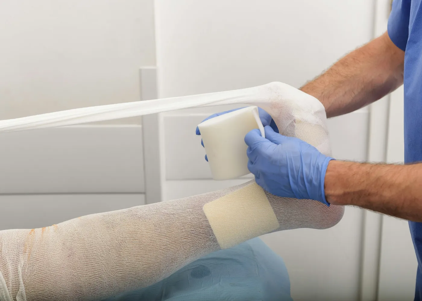 Medical professional applies a white compression bandage to a patient's leg with protective blue gloves in a clinical setting.