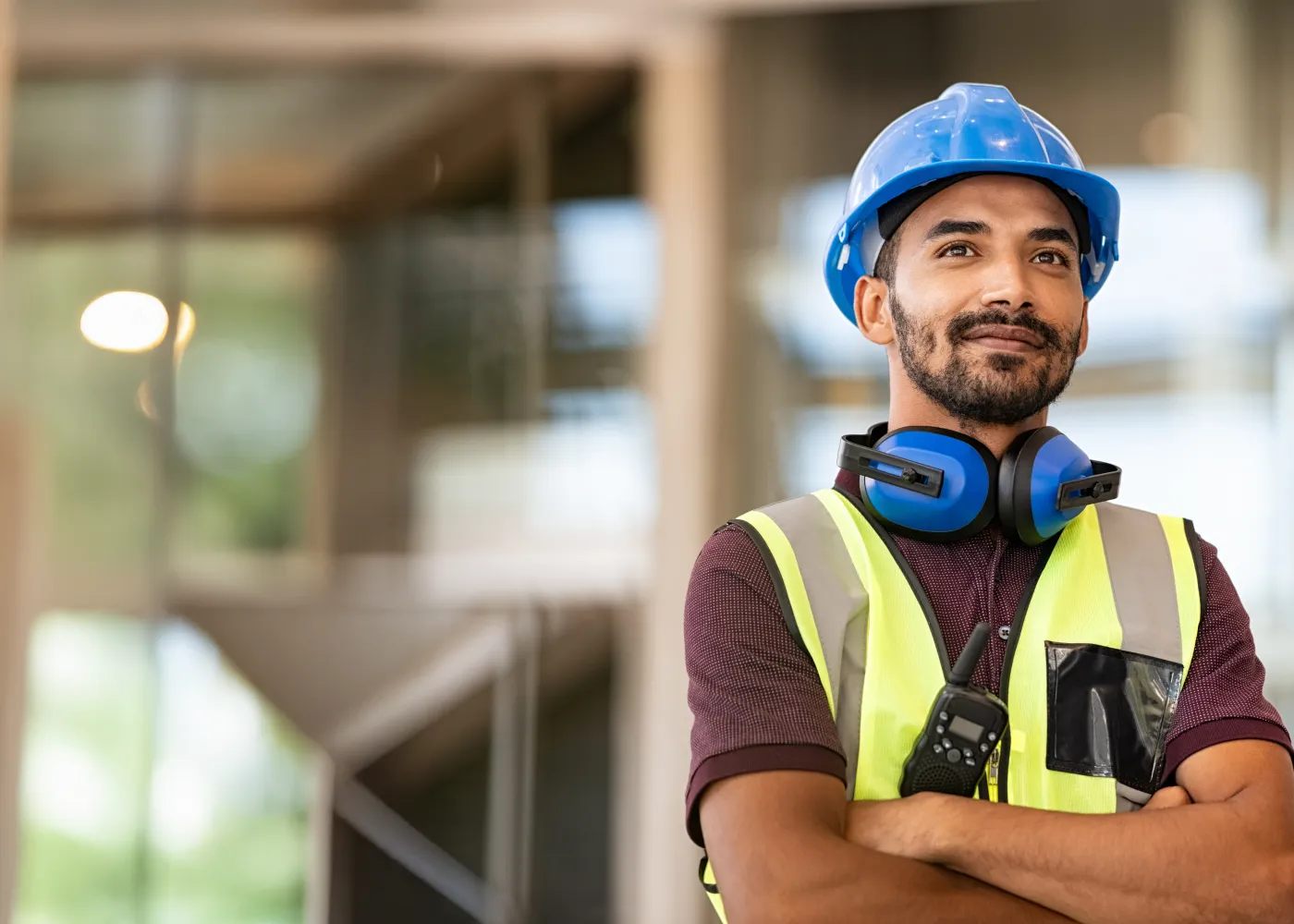 A construction worker in safety gear, blue helmet, and headphones, smiling confidently at the worksite.