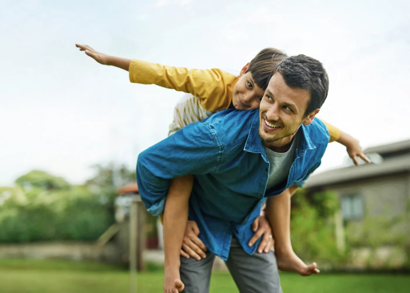 Father giving piggyback ride to son outdoors with greenery and clear sky in background