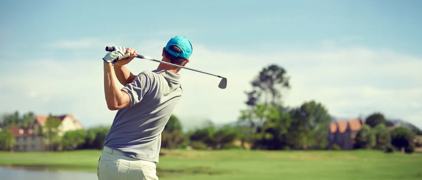 Male Golfer wearing a blue cap swings a club on a sunny day at a green golf course with distant houses.