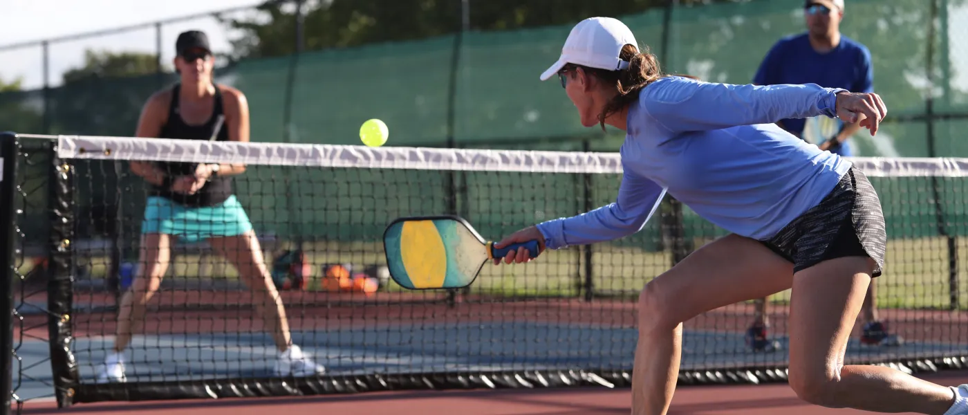 Middle aged woman lunging to hit a pickleball with a paddle on an outdoor court during a game.