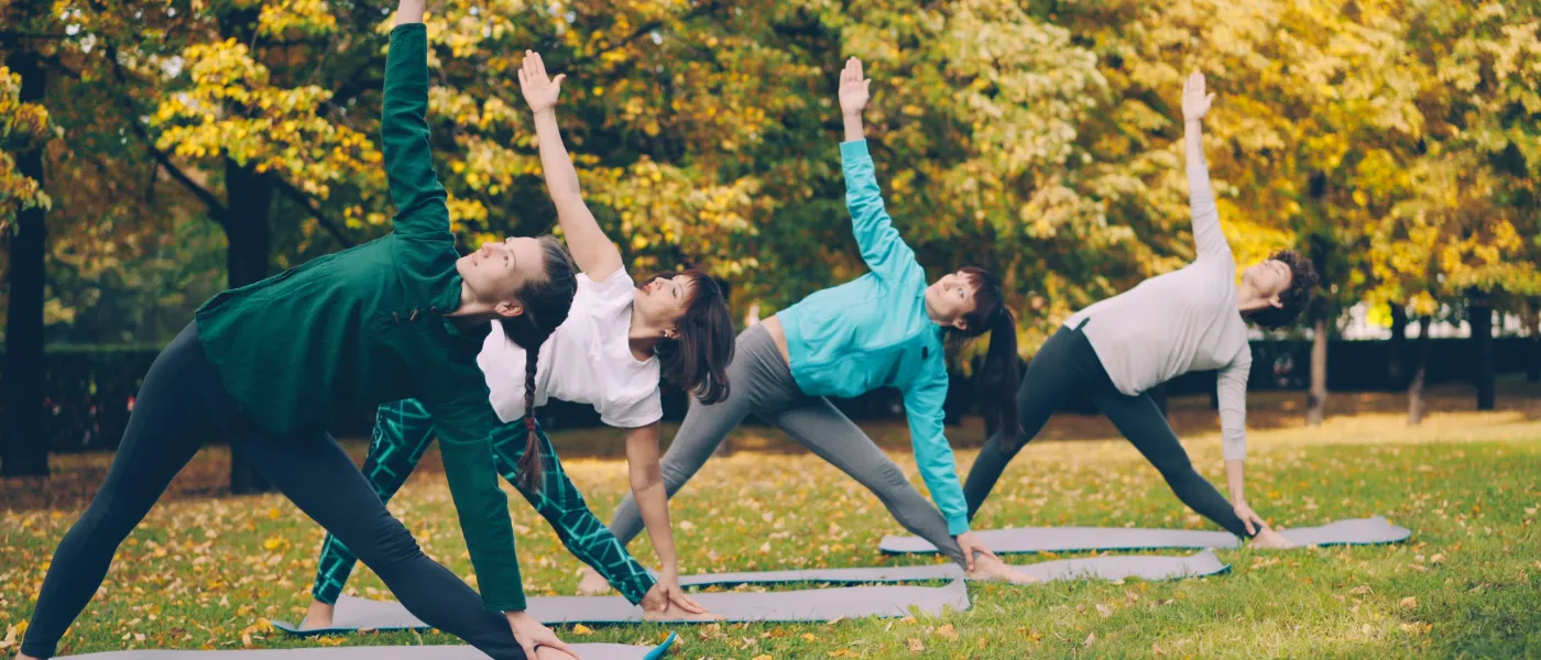 Four women practicing outdoor yoga in triangle pose on mats amid autumn foliage in a park.