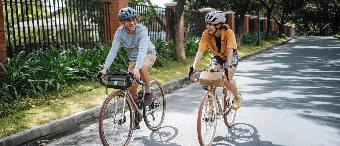 Two people wearing helmets smiling and riding bicycles on a sunny suburban street with greenery and houses.