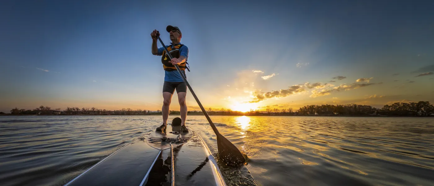 One Man paddleboarding on calm water during a vibrant sunset with clear sky and distant trees.