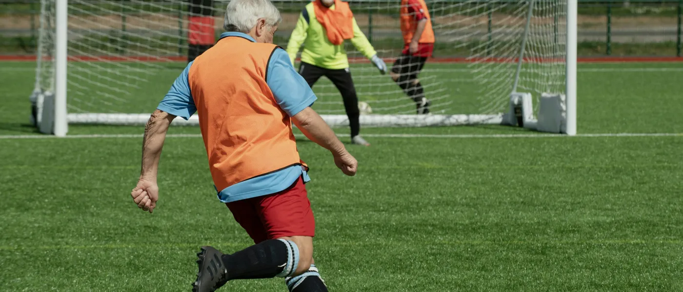 Senior man wearing orange vest kicking soccer ball towards goal with goalkeeper and defender ready to block