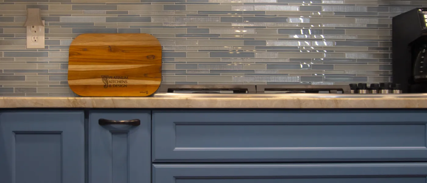 Modern kitchen countertop with blue cabinets, gray tile backsplash, wooden cutting board, and stovetop.