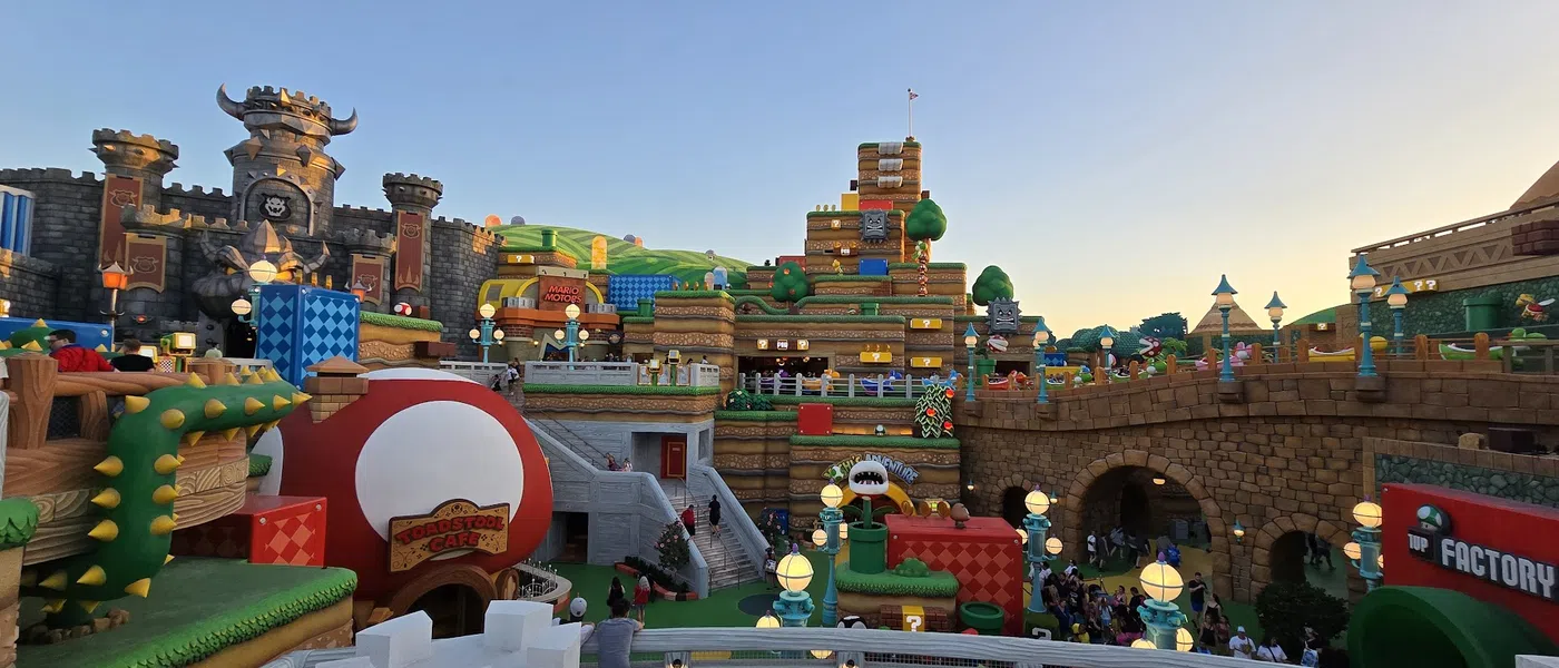 Colorful Super Mario-themed amusement park area with castle, giant toad house, and themed structures under clear sky.