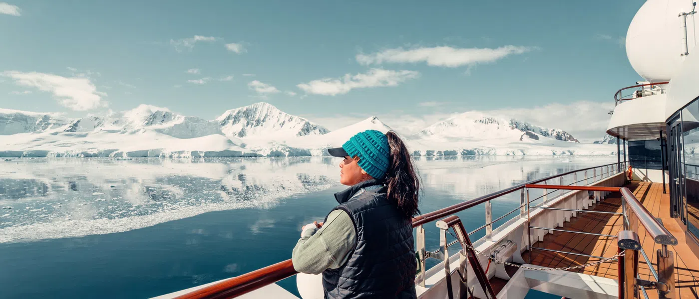 Woman in warm clothing gazing at snowy mountain range reflected on calm icy waters from a ship deck under clear blue sky.