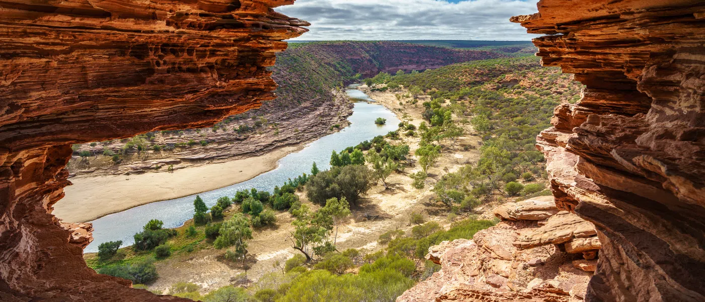 View through rocky cave opening overlooking winding river, greenery, and rugged cliffs under cloudy sky.