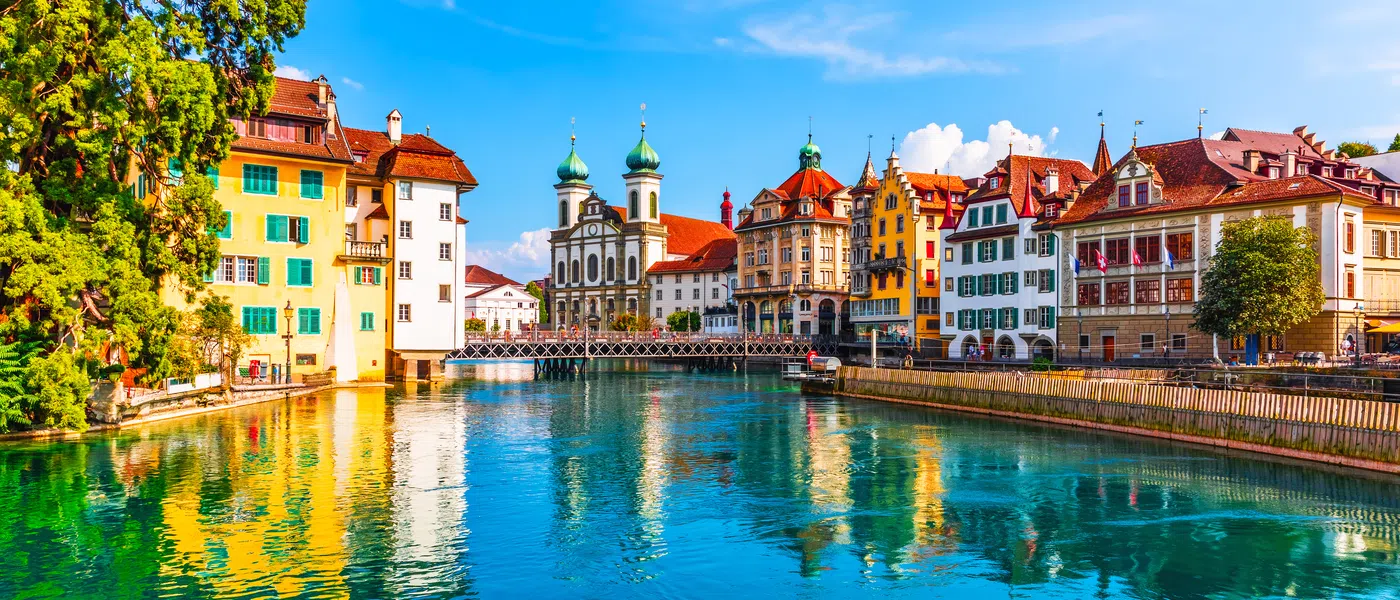 Colorful historic buildings and church reflect on the Reuss River under a bright blue sky in Lucerne, Switzerland