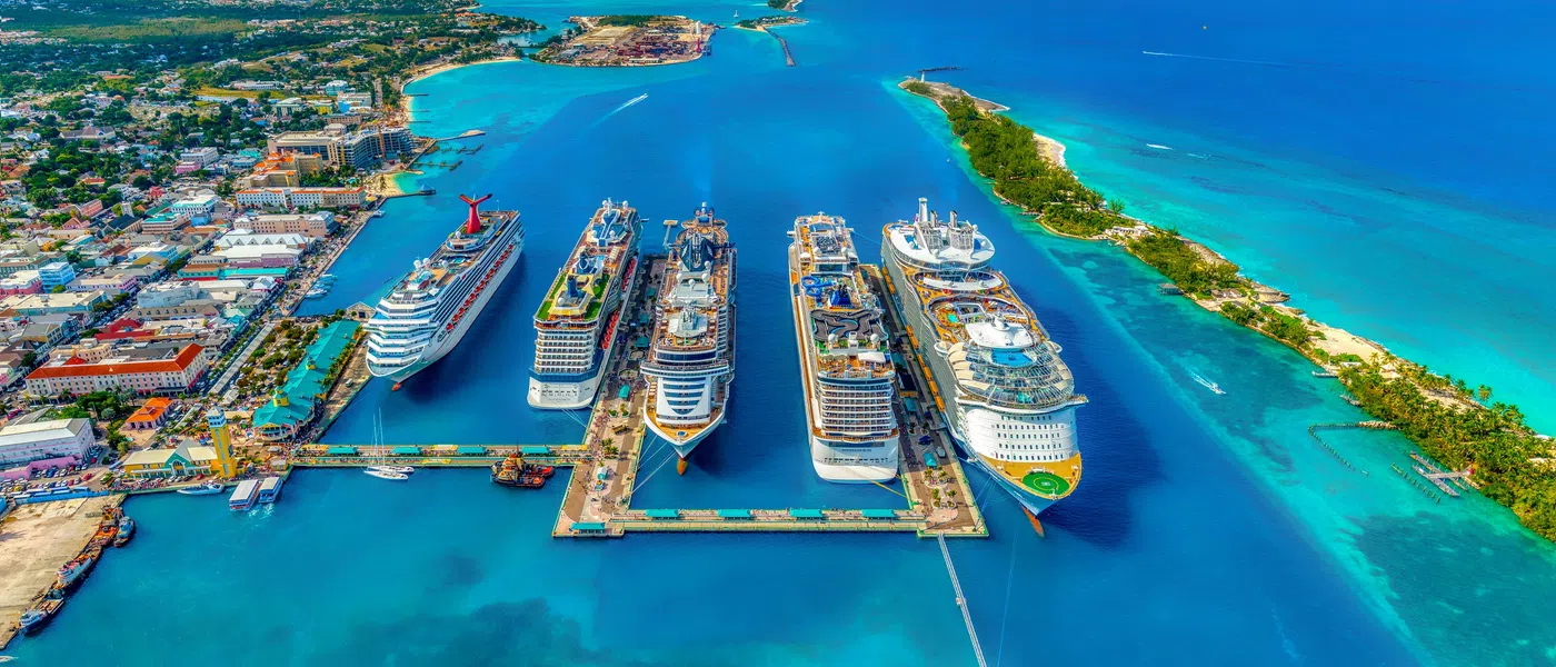 Aerial view of a busy cruise ship port with six large ships docked and turquoise ocean surrounding them