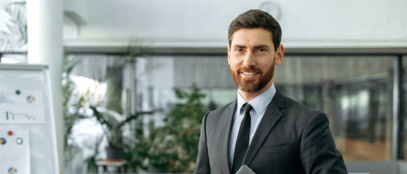 Smiling businessman in black suit holding laptop in modern office with bookshelves and flip chart