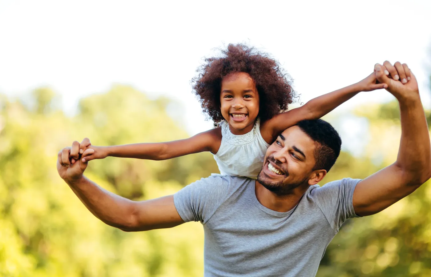 Smiling father holding daughter on his shoulders outdoors with trees in the background on a sunny day