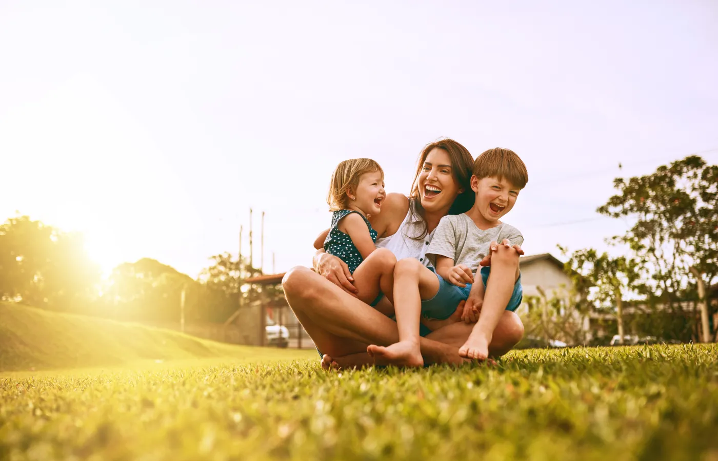 Happy mother playing and laughing with her two young children outdoors on grass during sunset.