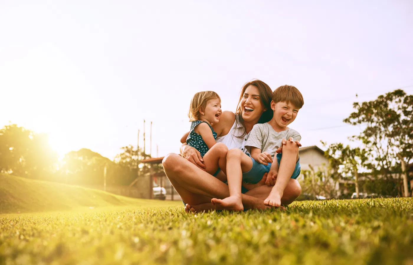 Mother sitting on grass outdoors laughing with two young children during golden hour in a park.