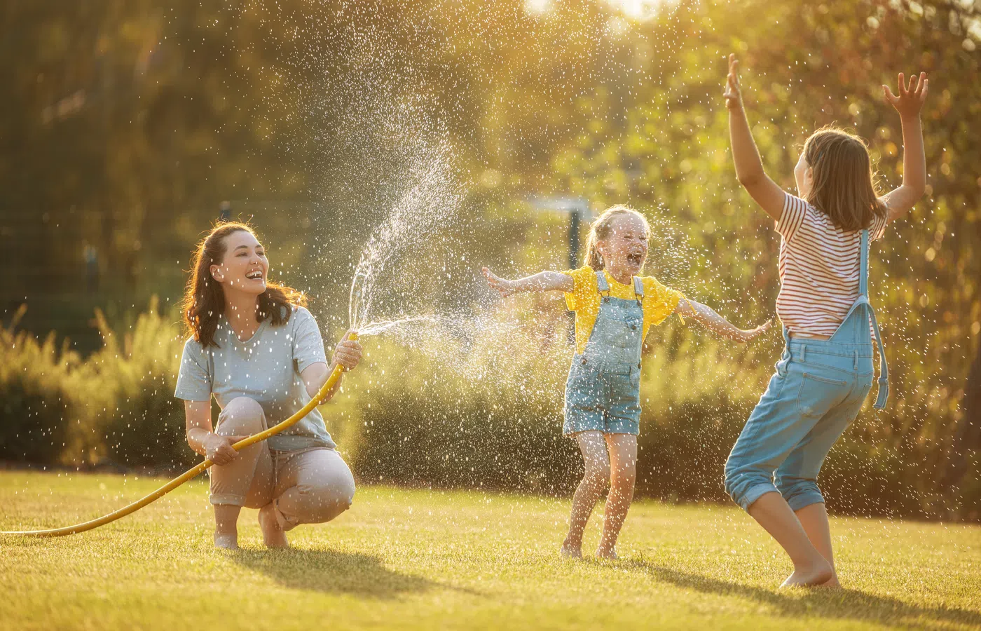 Mother sprays water with hose on two joyful children playing in sunny backyard on warm day