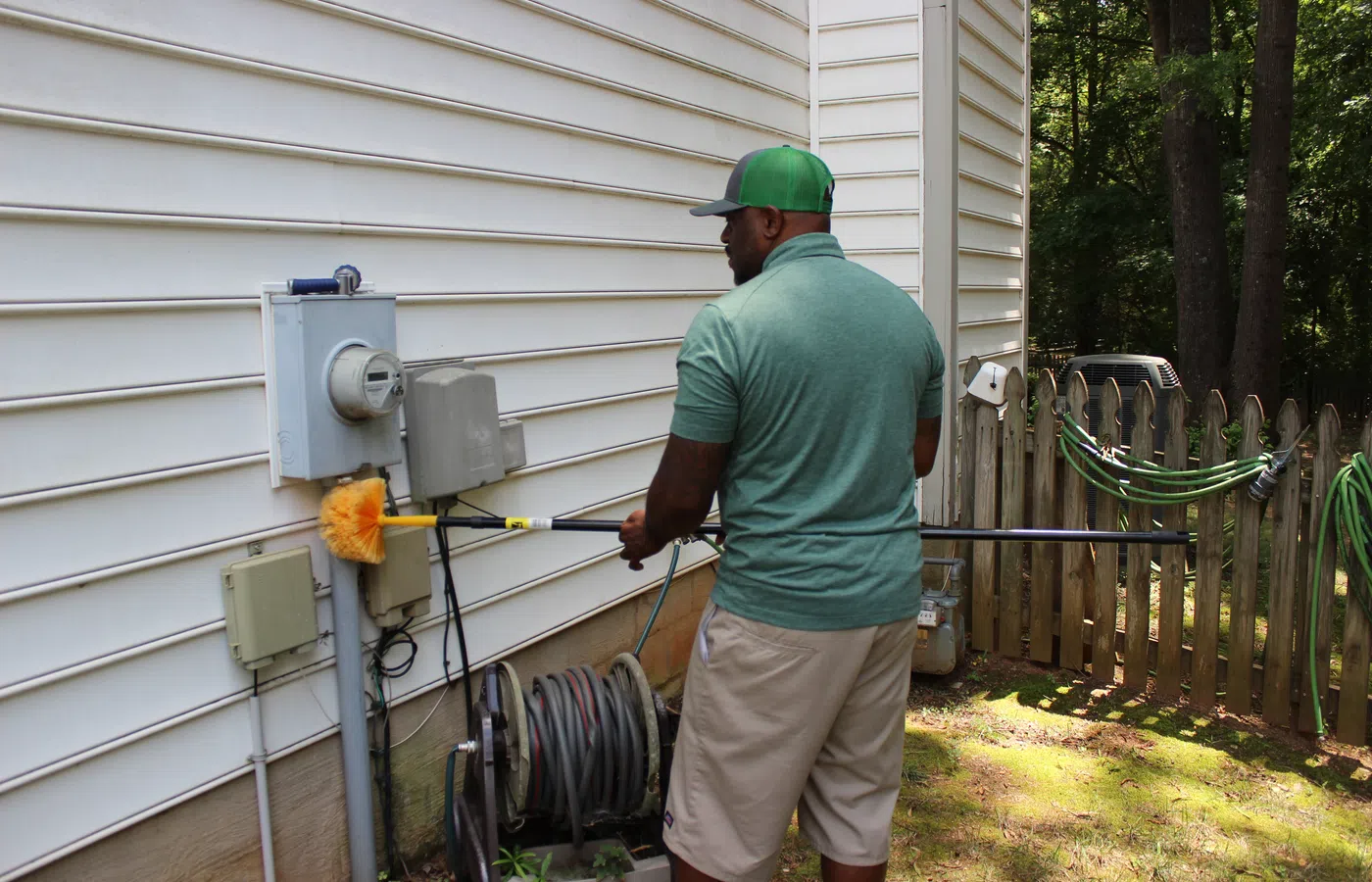 Man cleaning electrical meter and panel with a long brush outside a white house on a sunny day.