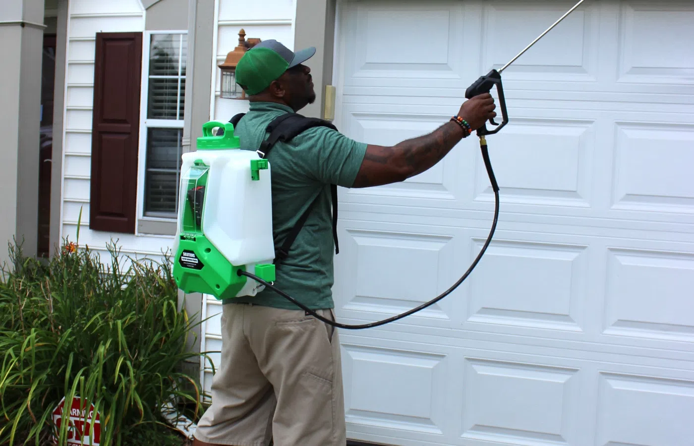 Man in green shirt and cap using backpack sprayer to spray white garage door outdoors.