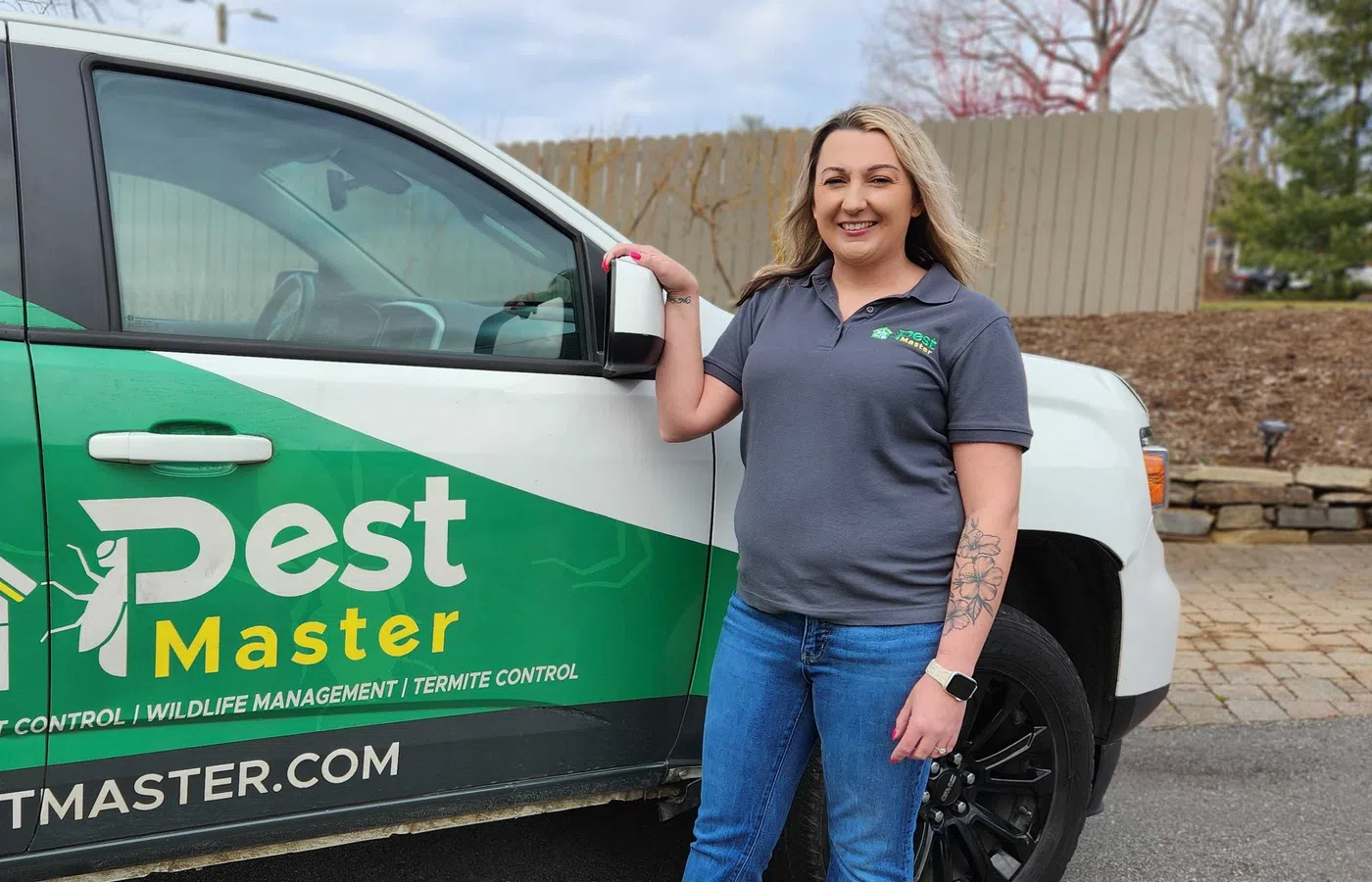 A woman in casual workwear stands beside a green and white Pest Master pest control vehicle outdoors.