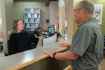 Smiling receptionist talking to a senior man at medical office reception desk with a co-worker in the background.