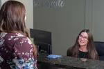 Receptionist smiling and assisting a woman at a clinic front desk with computer and sign behind.