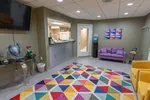 Colorful dental office waiting room with purple sofa, yellow chairs, geometric rug, and TV mounted on wall.