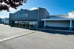 Gray storefront of At Home store with large windows and yellow bollards under a clear blue sky