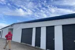 Person in red hoodie pressure washing white storage unit building with black doors under a blue cloudy sky.