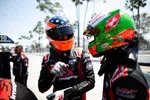 Two race car drivers in colorful helmets and racing suits sharing a moment at the pit stop on a sunny day.